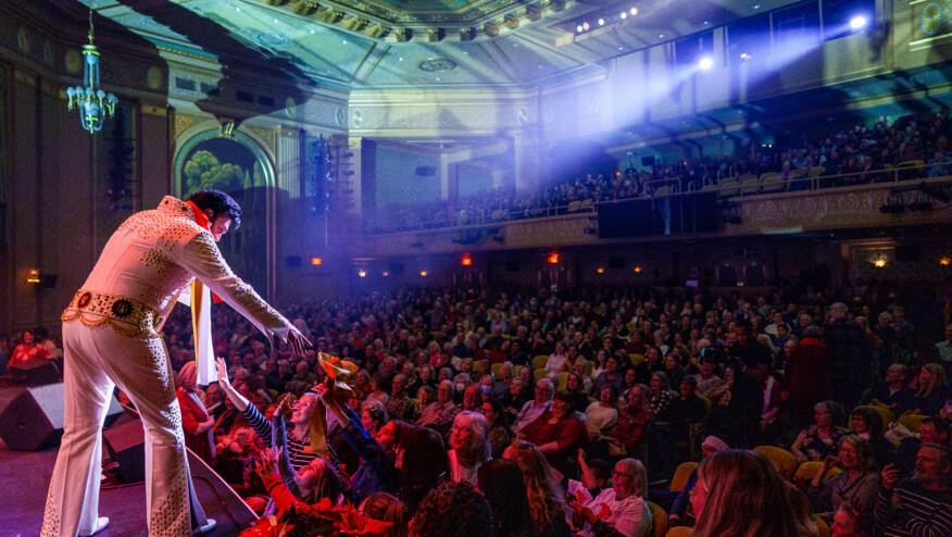 A man in a white jumpsuit stands onstage, reaching into the audience.