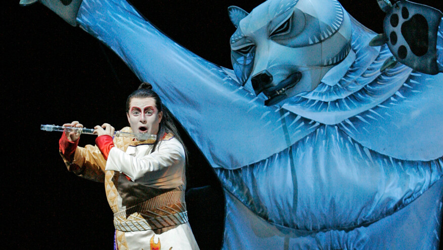 A large cardboard cutout of a bear looms behind a man playing a glass flute.