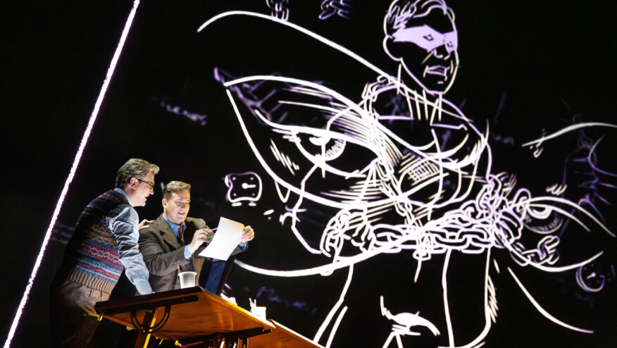 Two men stand at a table, reading a piece of paper. A drawing of a superhero is behind them.