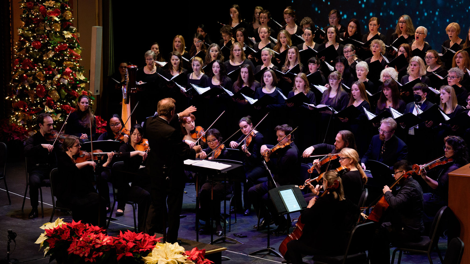 A man conducts musicians on a stage with flowers.