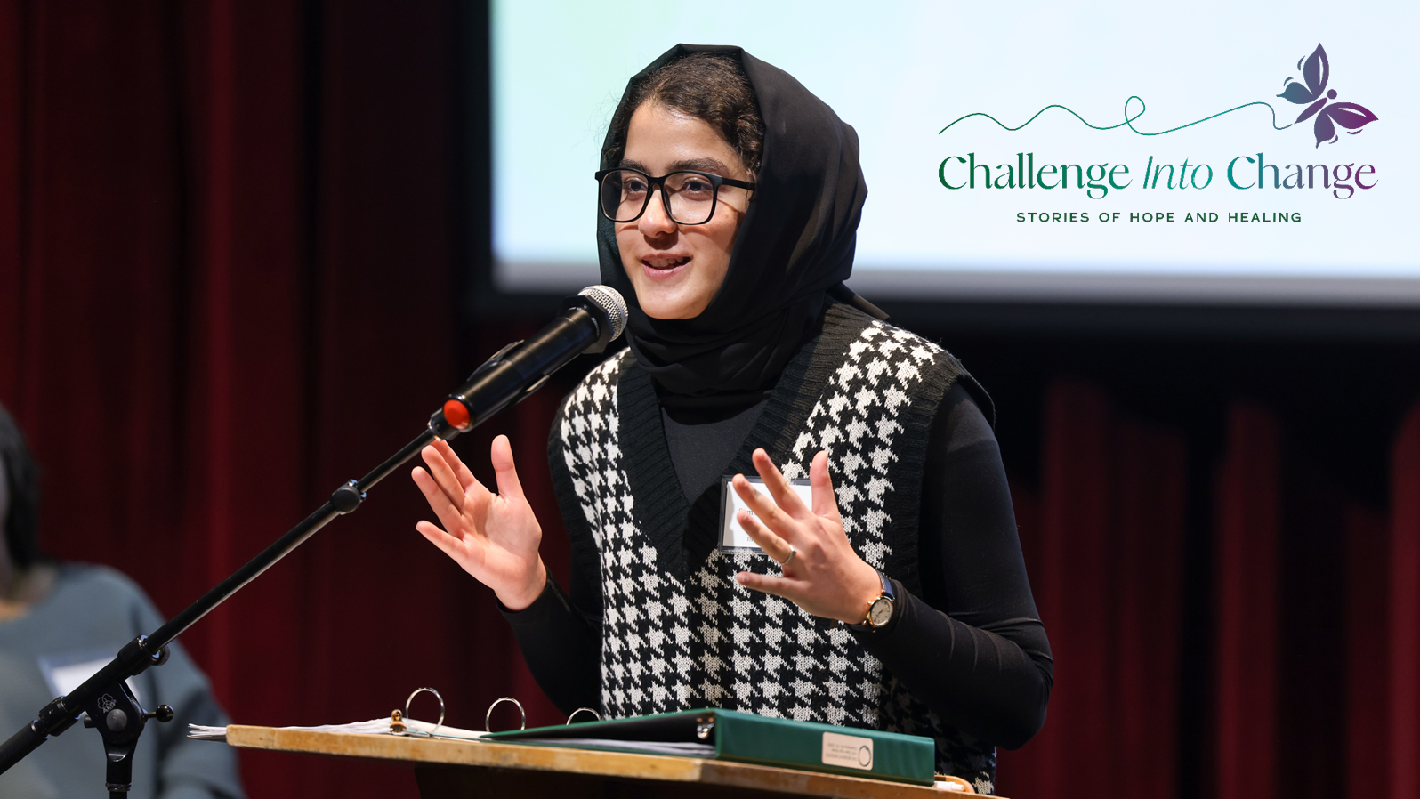 A young woman speaks at a podium on a stage. Text reads, 